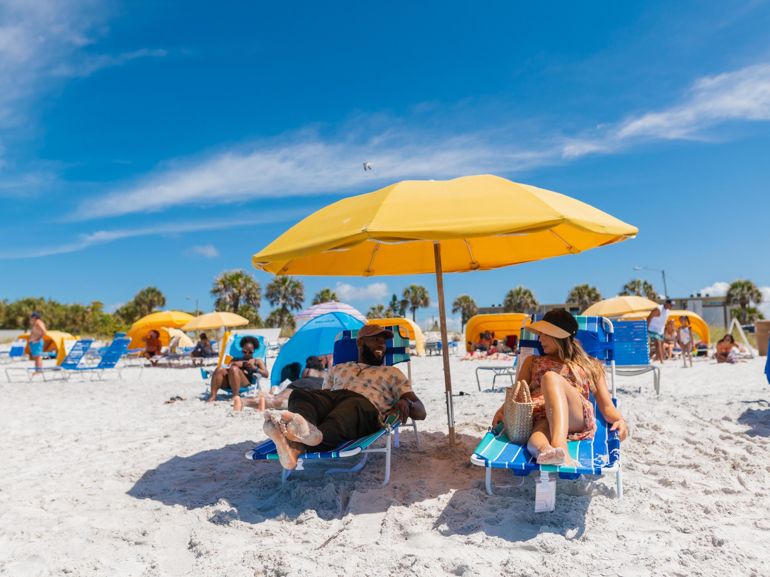 a couple relaxes on lounge chairs under a bright yellow umbrella on the white sands of Madeira Beach