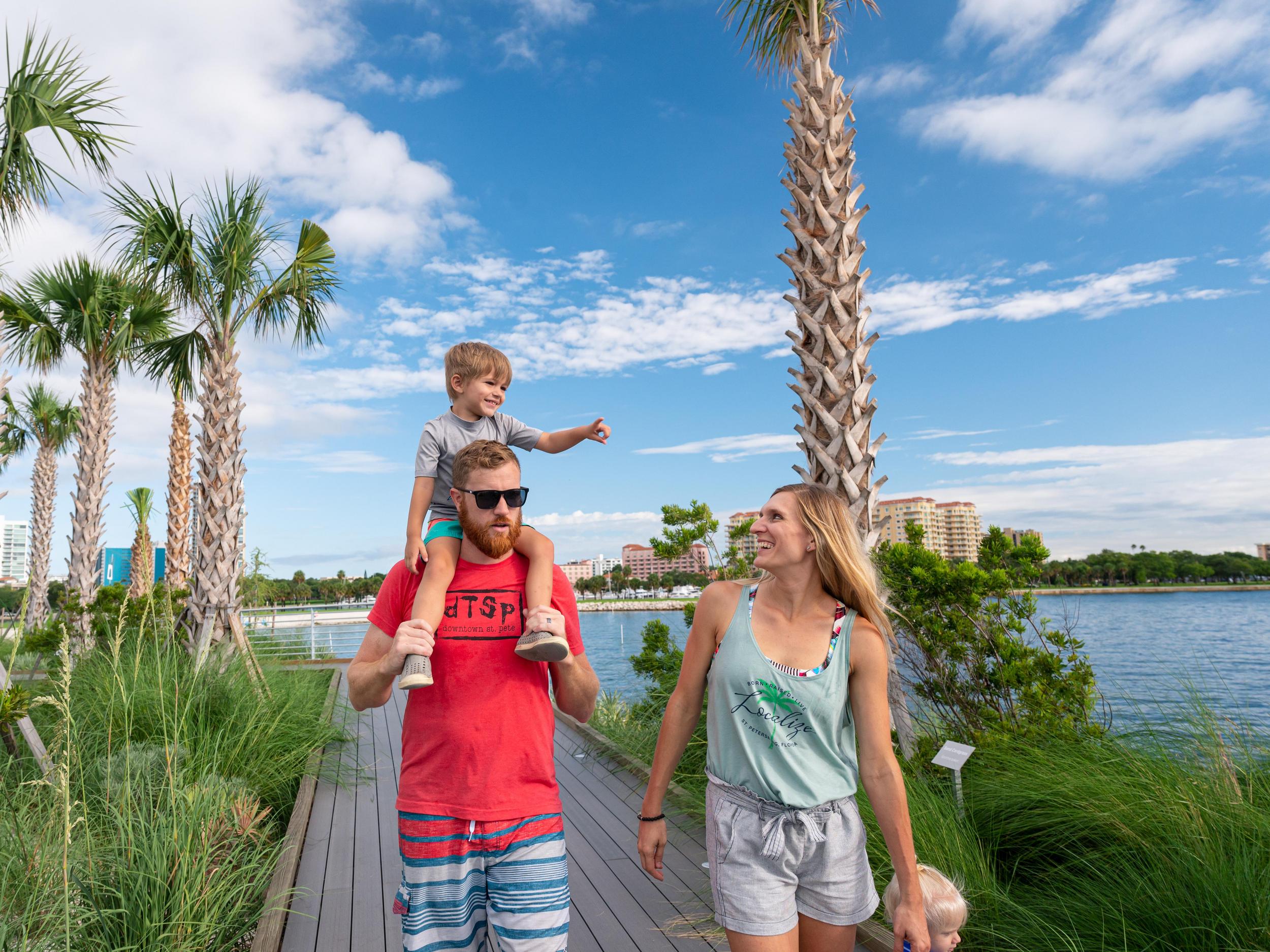 father with little boy on his shoulders walking next to a woman on a boardwalk surrounded by palm trees near the water