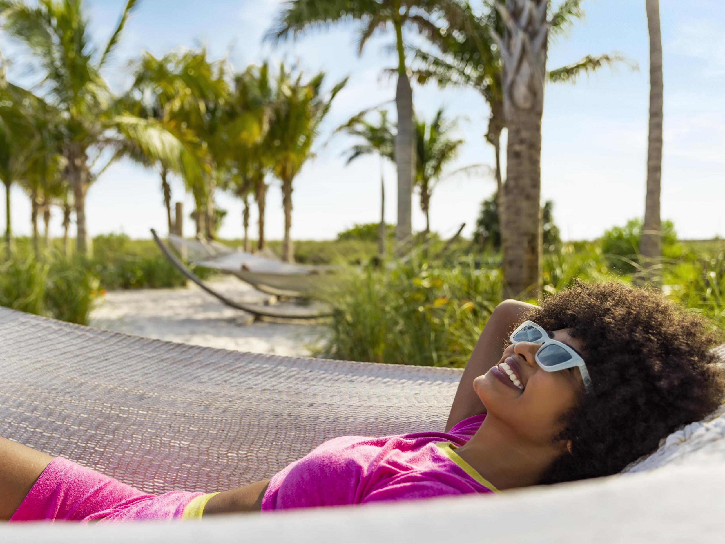 Woman with sunglasses laying in a hammock with the beach and palm trees in the background