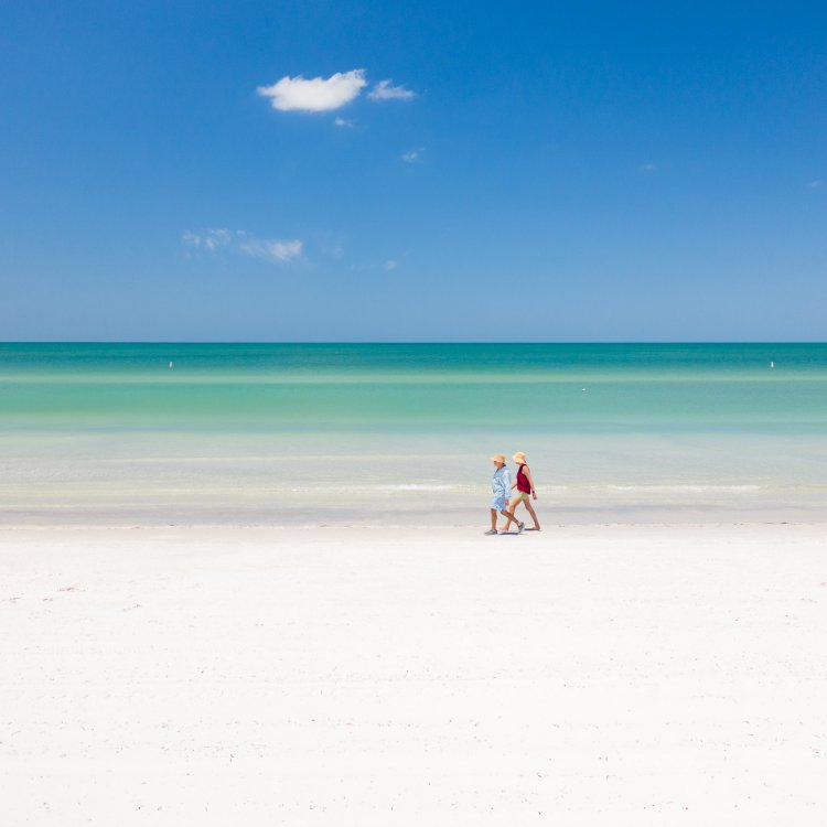 Two beachgoers walk along the white sandy shoreline and emerald green waters of St. Pete Beach on a bright sunny day.