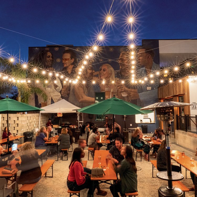 people drinking beer at long picnic tables outside with twinkling lights and a mural in the background at Grand Central Brewhouse