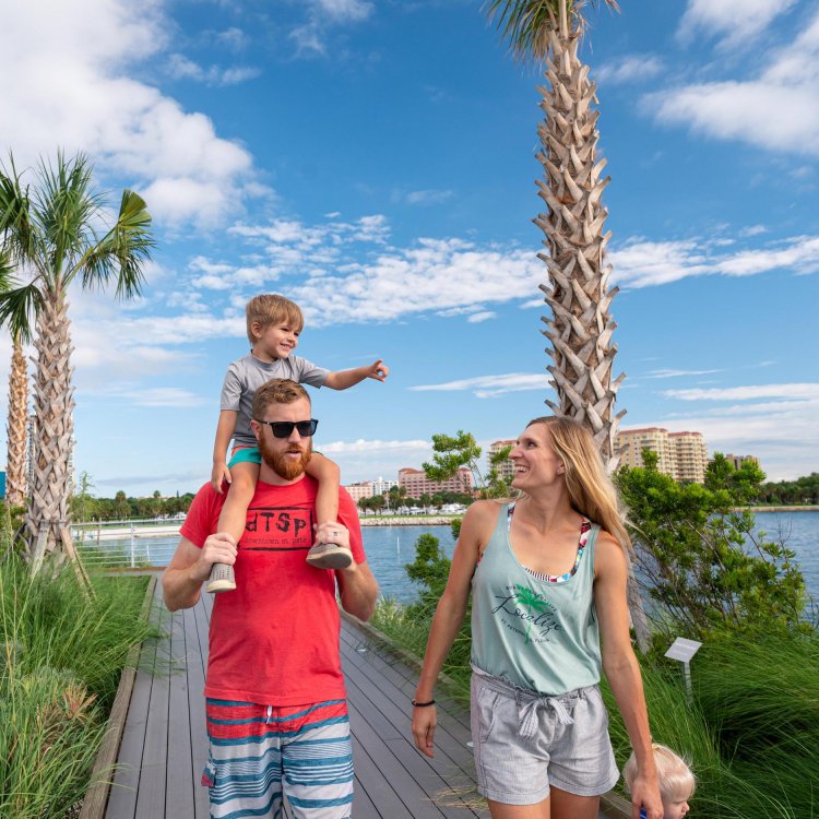 father with little boy on his shoulders walking next to a woman on a boardwalk surrounded by palm trees near the water