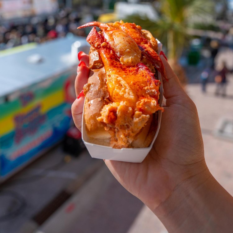 a hand holding a lobster roll stuffed with fresh lobster in a paper container with the John's Pass Seafood Festival in the background