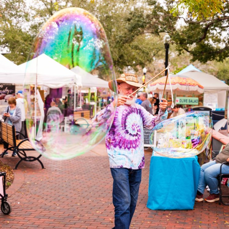 Man in a tie dye shirt blowing big bubbles at an outside market space.