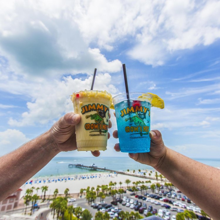 two tropical drink in a Jimmys Crow's Nest plastic cup cheersing with rooftop view in background of Clearwater Beach and Pier