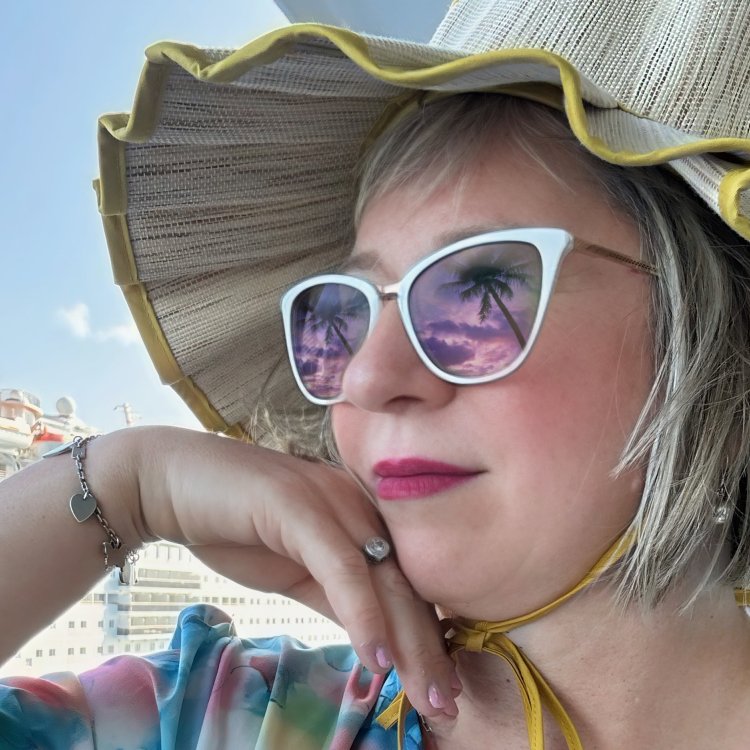 a close-up shot of a woman's face with white sunglasses that are reflecting palm trees and a beach scene; she is wearing a ruffle-edged straw hat and a multi-colored top