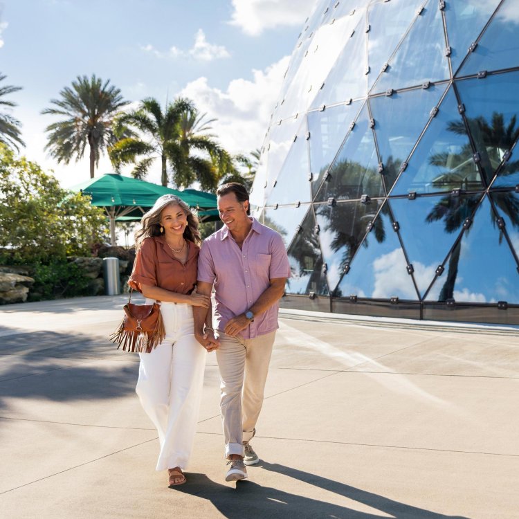a smiling mature couple walks outside the Dali Museum, where palm trees are reflected in the mirrored building exterior called "The Enigma"