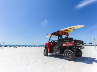 Am Strand parkt ein Rettungsschwimmer-Buggy mit einem Surfbrett darauf. In der Ferne sind Cabanas zu sehen.