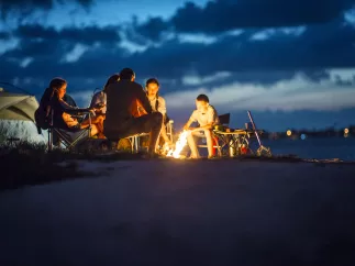A family circles a bon fire on the beach with a tent in the background