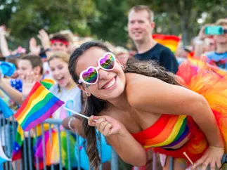 a happy lady dressed in colorful outfit wearing colorful sunglasses holding a LGBTQ+ flag stands out from the crowd during Pride parade.