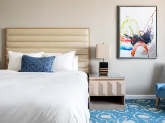 Showing the inside of a hotel room, queen size bed, blue and white carpet, nightstand and abstract painting on the wall. 
