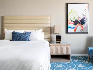 Showing the inside of a hotel room, queen size bed, blue and white carpet, nightstand and abstract painting on the wall. 