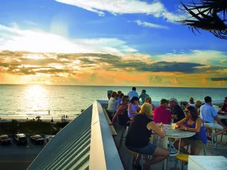 Roof top bar with people enjoying drinks and an ocean view with a sunset in background. 
