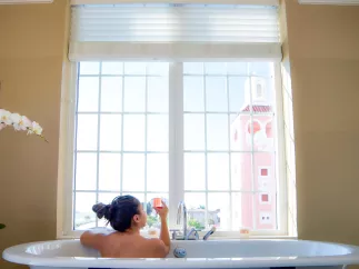 A woman in a bath tub holds a pink champagne glass while looking out the window at the Don Cesar at Spa Oceana