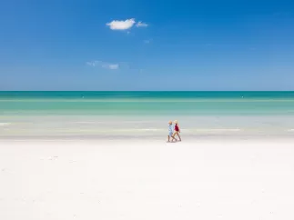 Two beachgoers walk along the white sandy shoreline and emerald green waters of St. Pete Beach on a bright sunny day.