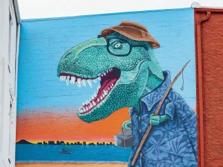 a mural depicts a tyrannosaurus rex dressed as if he is going fishing with a fishing pole, hat and tackle box. In the water behind him, a smaller dinosaur rides a jet ski