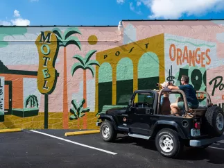 A man and woman in a black jeep park in front of a vintage-themed mural with signs for "motel," "oranges" and "all are welcome here"