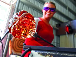 A woman holds an orange piece of art glass in a hot shop