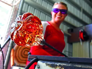 A woman holds an orange piece of art glass in a hot shop