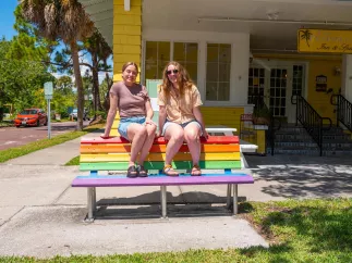 two women in shorts and tshirts sit on top of a bench painted in rainbow colors in front of a bright yellow inn next to a street