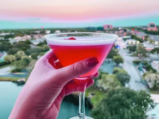 a manicured hand holding a pink cocktail with a cherry with a view of sunset and the Gulf from Vantage Rooftop Bar at the Karol Hotel