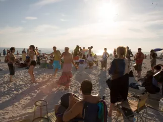 A group of beach-goers dance in the sand to drums on the beach at sunset.