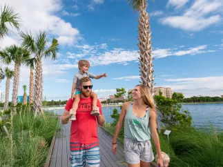 father with little boy on his shoulders walking next to a woman on a boardwalk surrounded by palm trees near the water