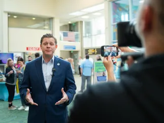 a man in a business suit at the airport giving a speech that is being recorded by a person holding a small video camera
