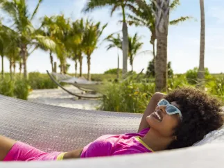 Woman with sunglasses laying in a hammock with the beach and palm trees in the background