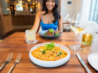 a smiling woman in a bright blue dress sits across from a bowl of pasta at a restaurant