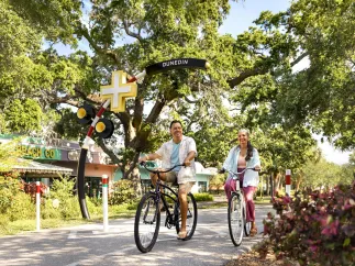 a middle-aged couple rides bikes on the Pinellas Trail in Dunedin under the cover of live oak trees, with businesses nearby