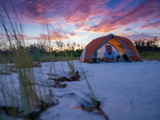a young man relaxes inside his tent on the beach at Shell key with a dramatic pink and blue sky behind him