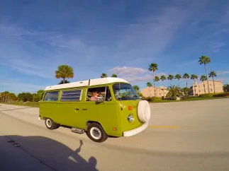 a vintage green van driving on a road with buildings and palm trees in the background