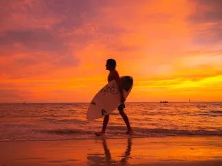 a man with a surfboard walks along the water's edge in Treasure Island at sunset