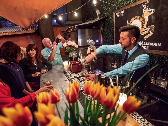 Group of people sitting at a bar with a bartender pouring drinks. Foreground has a vase full of lilies