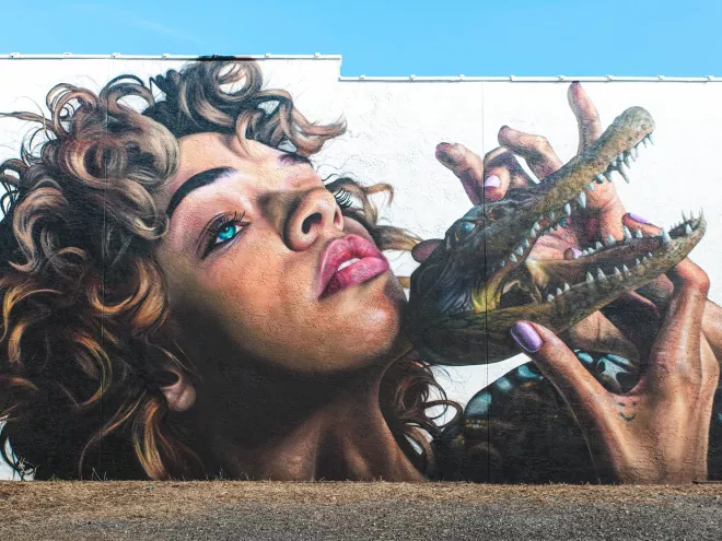 The "Mask Off" mural by Drew Merritt in St. Pete depicts the head of a beautiful Black woman with curly hair and blue eyes holding a small alligator in front of her