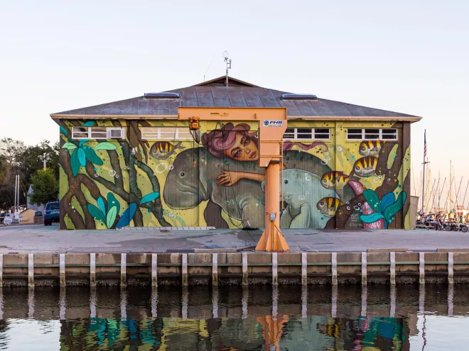 a mural on the side of a building depicts a woman with swirling reddish hair riding or hugging a manatee while her calf and fish swim nearby