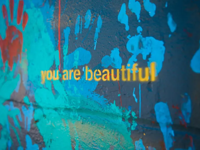 a portion of a large mural displays the words "you are beautiful' in yellow lowercase letters against a background of painted blue, green and red handprints on a dark background