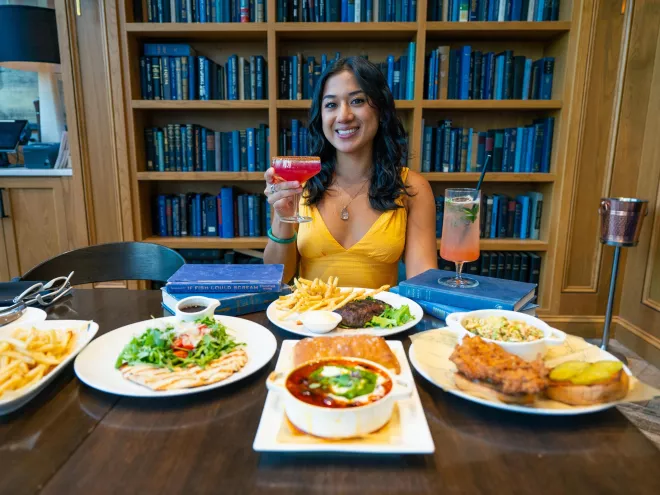 A woman in a yellow dress holds up a cocktail in front of plates of food with a bookshelf behind her