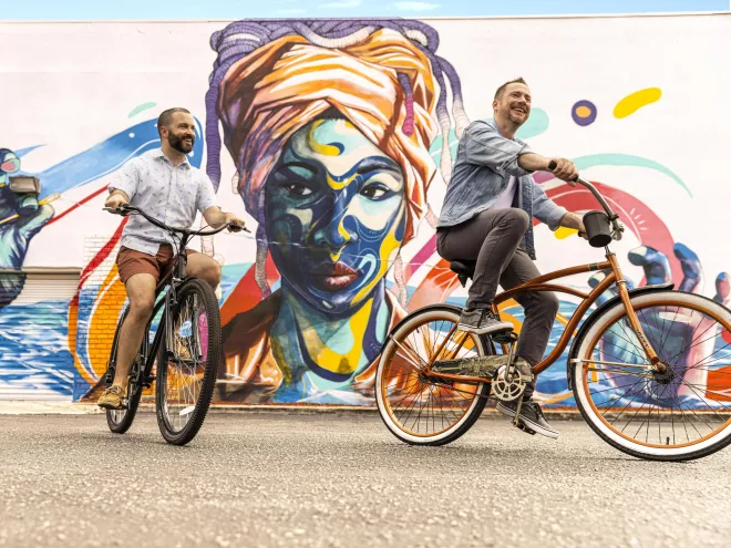 two men riding bikes in front of a mural of an African woman with a turban