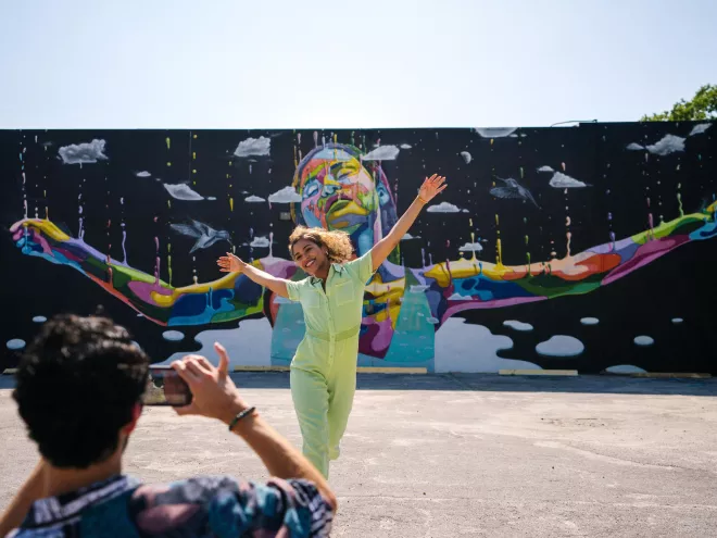 a person taking a photo of a woman wearing a lime green jumpsuit with her arms outstretched in front of a mural of a black woman with her arms outstretched