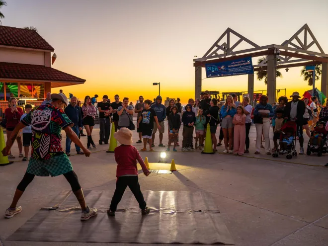 a performer talks with a young boy across from a gathering of people with the sun setting behind them at Pier 60 in Clearwater Beach