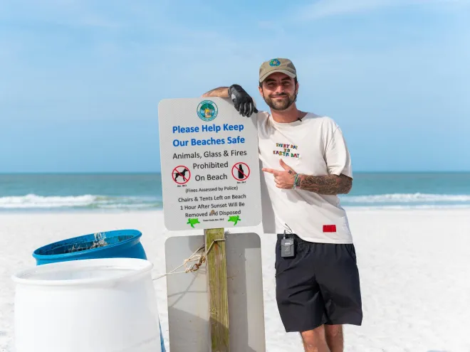 a young man standing next to a trash can and a sign at the beach encouraging people to pick up trash