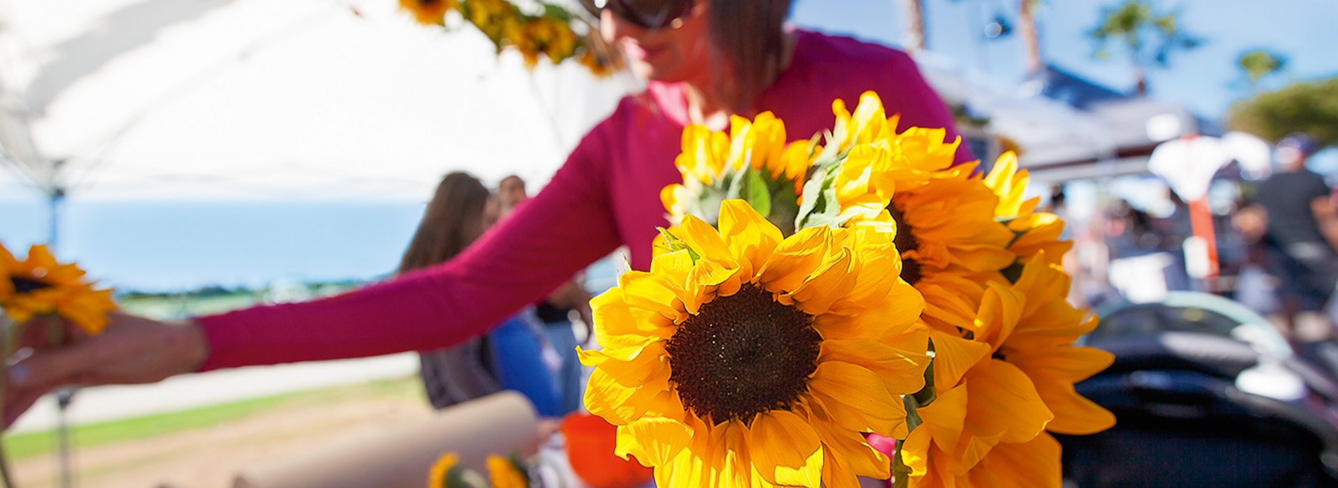 A woman holds a bundle of sunflowers at a sunny, open-air market.