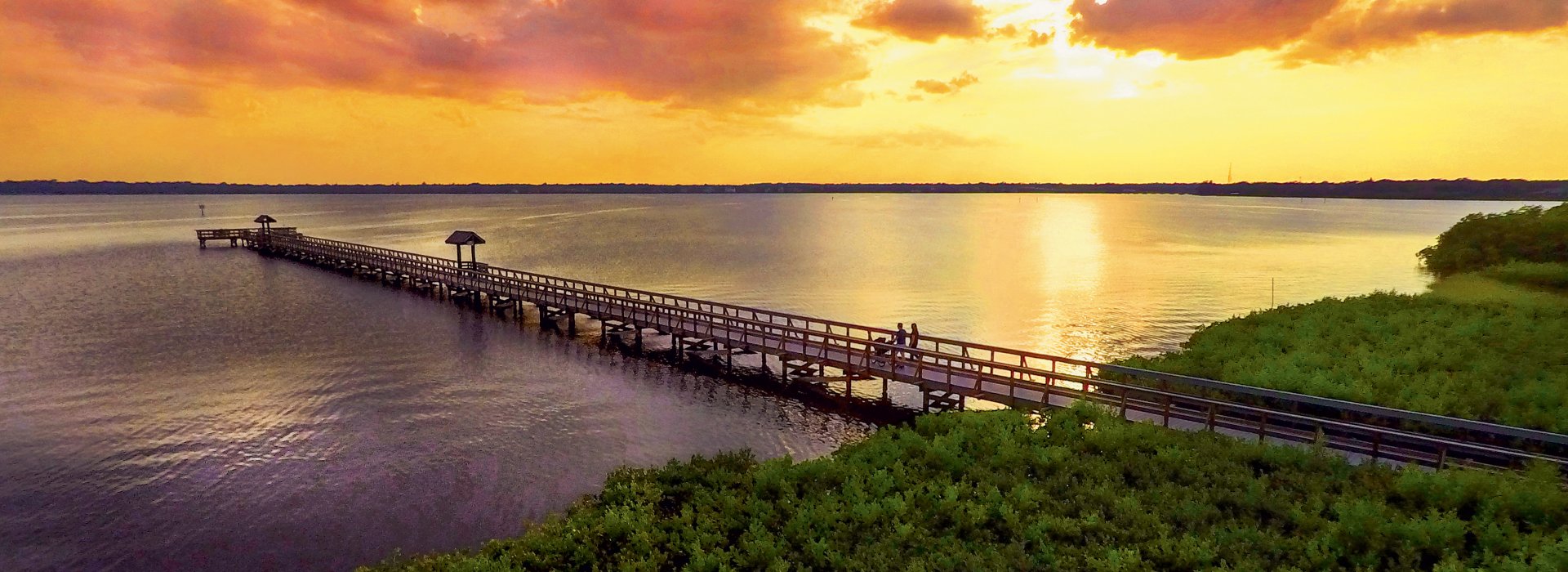 Aerial view of the pier at R.E. Olds Park in Oldsmar, with lush greenery on land and a brilliant orange sunset