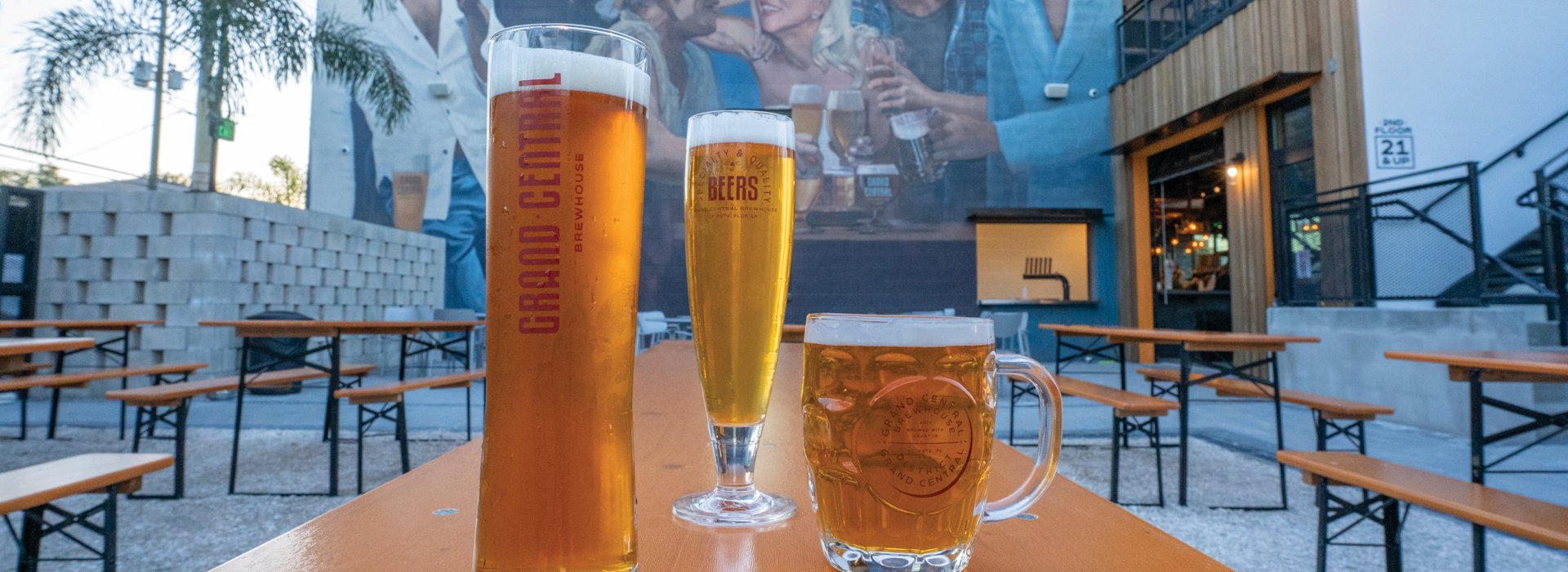 Three glasses of beer on top of an outside table in front of a large mural at the Grand Central Brewery in St. Pete.