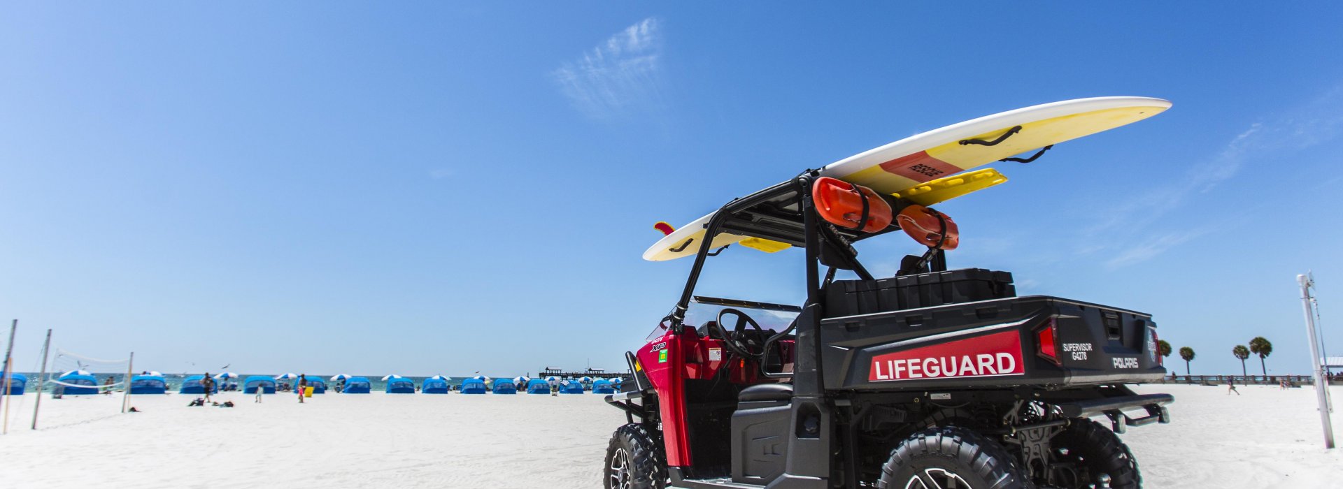 A lifeguard buggy parked on the beach with a surfboard on top. Cabanas can be seen at a distance.