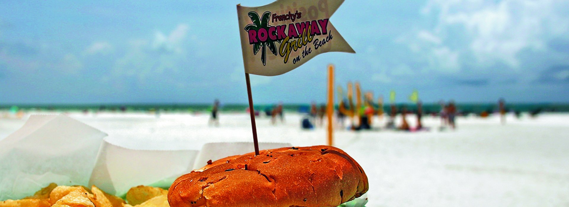 A grouper sandwich with fries at an outdoor patio with a view to the beach. Beach goers can be seen at a distance.