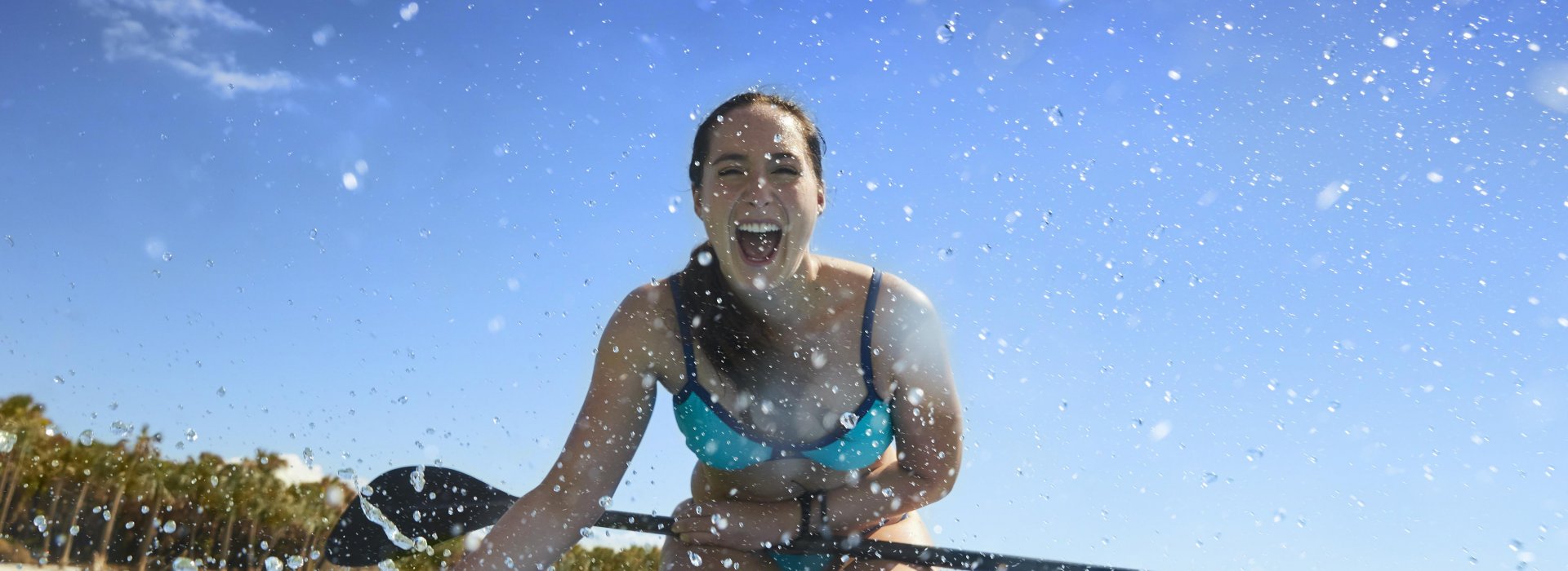 A girl splashing water at the camera while kneeling down on a stand up paddleboard.