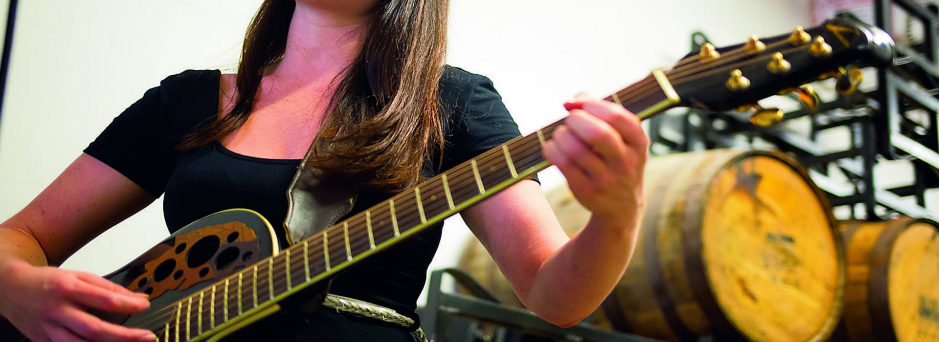 A lady playing a black guitar with beer barrels in the background.